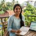 A 25-year-old woman in a light kurta sitting on a balcony, holding a notebook and a digital tablet showing financial growth charts.